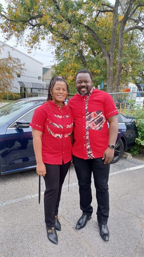 Two people wearing red shirts and black trousers and standing by a blue car.