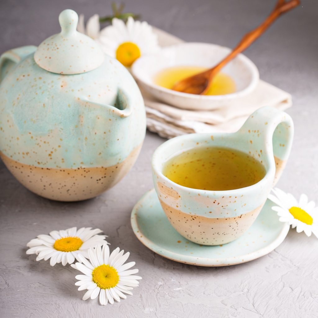 Chamomile tea in a tea cup, with a tea pot and bowl of honey on a white background.