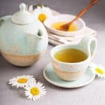 Chamomile tea in a tea cup, with a tea pot and bowl of honey on a white background.
