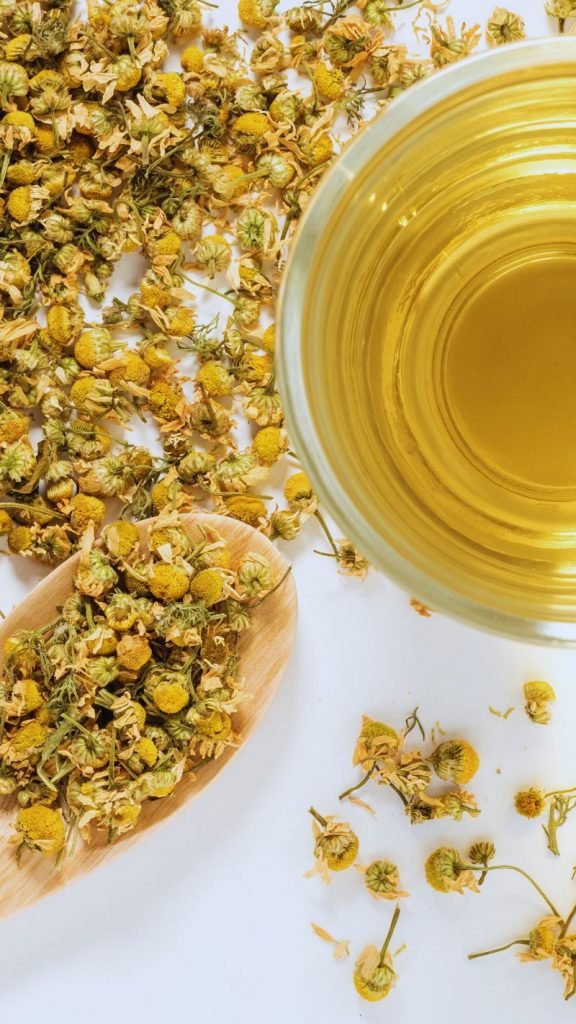 Chamomile tea in a glass tea cup, with dried chamomile flowers on a wooden spoon and white background.