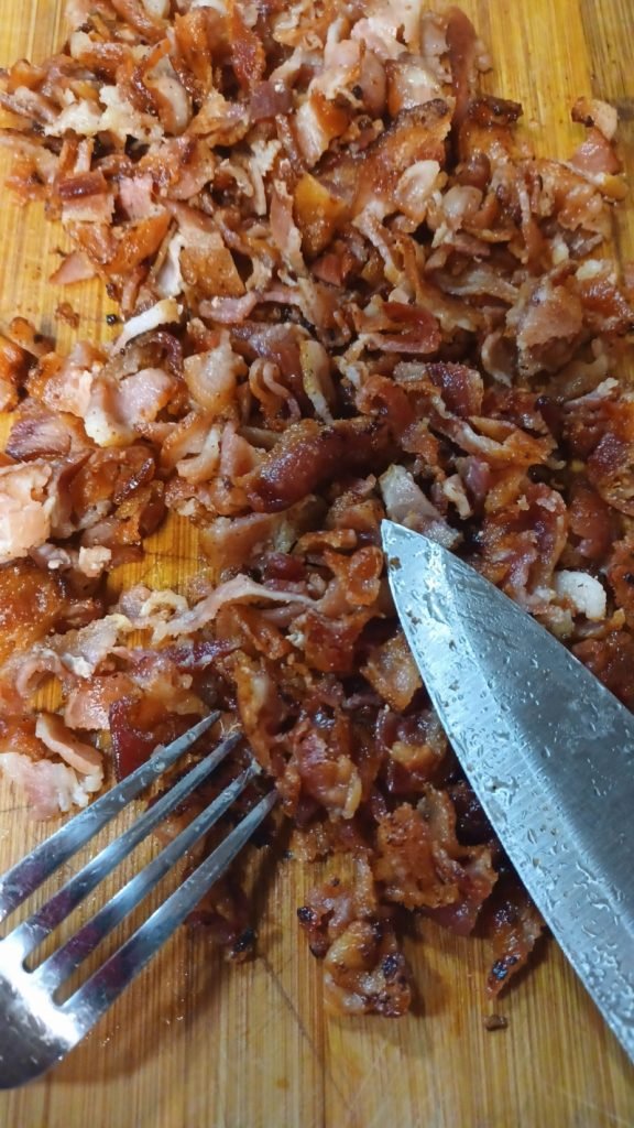 Chopped fried bacon on a brown chopping board with a fork and knife.
