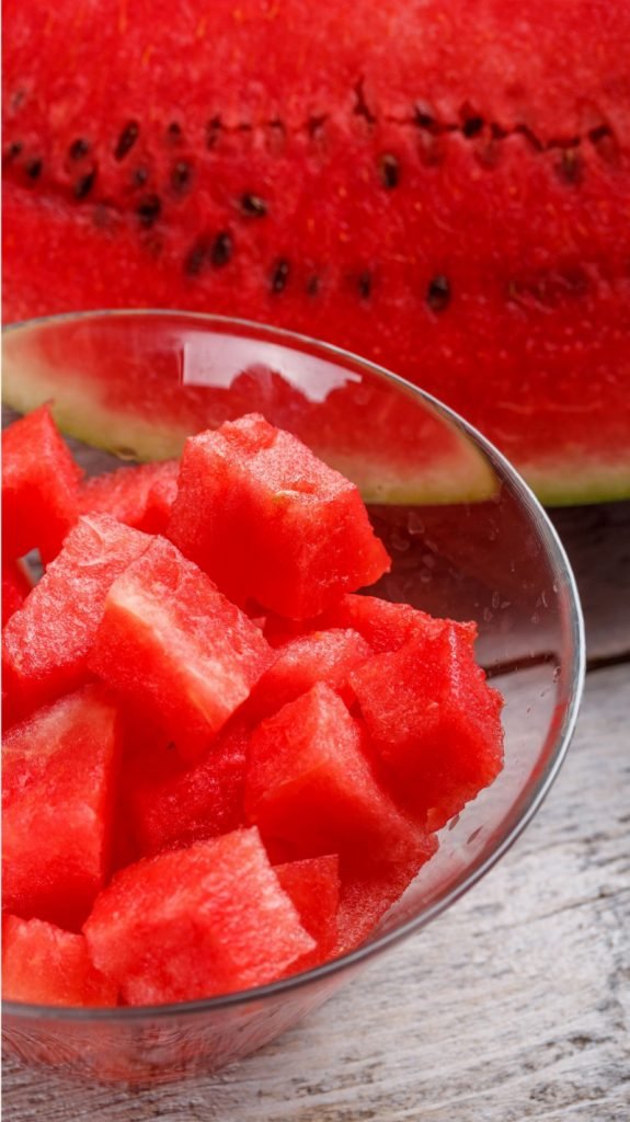 Diced watermelon in a glass bowl and a sliced watermelon piece behind it.