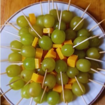 Green grapes and cheese stacked in brochettes in a white plate on a brown board.